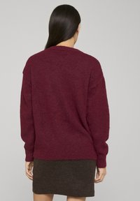 Pull en tricot bordeaux à coupe décontractée, épaules tombantes et ourlet côtelé. Texture douce, associé à une jupe ajustée marron foncé.