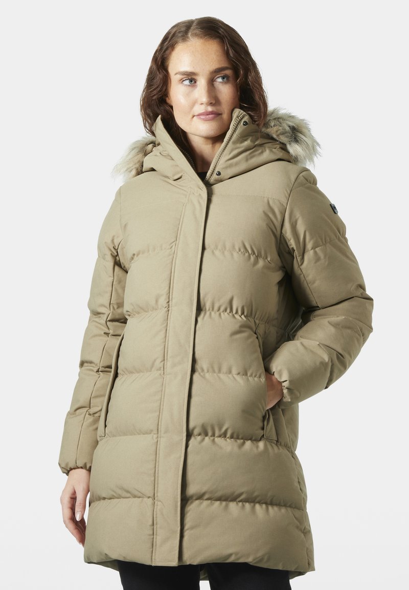 Mujer con abrigo invernal acolchado beige con capucha con borde de piel, de pie con una mano en el bolsillo, mirando hacia un lado sobre un fondo blanco.