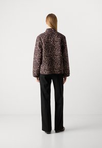 Person with long hair stands facing away, wearing a dark floral jacket, black wide-leg pants, and dark shoes on a white background.