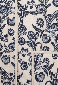 Υφασματάκι με ανάγλυφο μοτίβο, διακοσμημένο με ναυτικά λουλούδια σε εκρού φόντο, με ορατά ραψίματα, κούμπωμα με κουμπί και μικρή τσέπη.