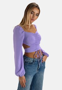 Lavendel langærmet top med rynket foran og udskæringer i siden, parret med lyseblå højtaljede jeans. Blødt, strækbart stof.