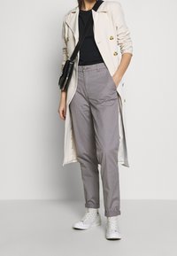 Lichtbeige lange trenchcoat met gouden knopen, gecombineerd met grijze tapered broek en witte sneakers. Schoon ontwerp met minimale accenten.