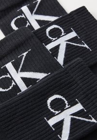 Black knitted socks with ribbed texture, featuring large white logo printed on the fabric. Three pairs displayed, overlapping slightly.