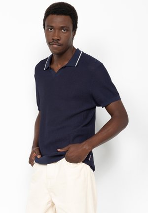 Homme portant un polo en maille bleu marine à manches courtes avec des bordures blanches, les mains dans les poches, assorti à un pantalon de couleur claire, debout devant un fond blanc.