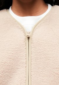 Jachetă din fleece cu un exterior texturat de culoare bej deschis, închidere cu fermoar de culoare cărămizie și un guler alb dedesubt. Material moale cu un aspect confortabil.