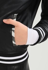 En hand vilar i fickan på en svart läderjacka och visar den randiga ribbade manschetten av en vit långärmad skjorta under.