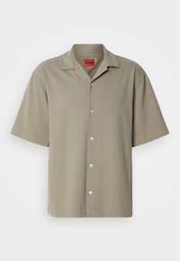 Chemise beige à manches courtes boutonnée avec col, affichant le nom de la marque "HUGO" sur une étiquette rouge au niveau du cou et un logo subtil sur la poitrine gauche.