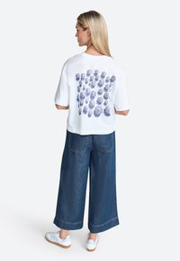 Weißes, kurzes T-Shirt mit einem blauen geometrischen Blumenmuster auf dem Rücken, kombiniert mit weit geschnittenen Jeanshosen und weißen Sneakers.