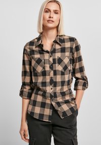 Chemise à carreaux noir et beige avec un devant à boutons, deux poches sur la poitrine et des manches retroussées, fabriquée en tissu doux avec une texture lisse.