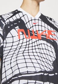 Koszulka Nike z czarno-białym, siatkowym wzorem, z wyraźnym czerwonym logotypem "NIKE" oraz małym czerwonym swooshem w lewym górnym rogu.