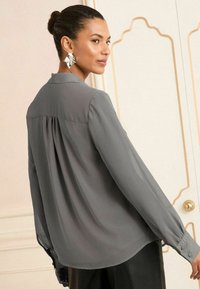 Bluză lungă, cu mâneci lungi, de culoare gri, cu manșetă cu nasturi, dintr-un material moale, cu detaliu plisat pe spate și guler clasic, purtată cu pantaloni negri.
