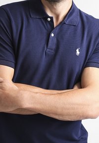 Man wearing a navy blue polo shirt with a white embroidered polo player logo, arms crossed across his chest.