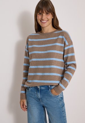 Smilende kvinde iført brun sweater med lyseblå vandrette striber og blå jeans, stående med den ene hånd i lommen mod ensfarvet baggrund.