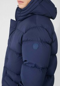 Personne portant une doudoune bleu marine avec un col montant et une capuche, comportant un logo circulaire sur le bras supérieur.