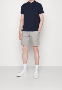 Polo shirt blu navy a maniche corte con patta a tre bottoni, abbinato a pantaloni corti grigio chiaro e sneakers bianche, esposto su uno sfondo neutro.