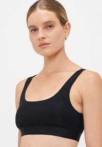 Young woman with sleek hair wearing a black seamless sports bra with wide straps and subtle logo on the band, neutral background.