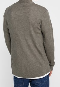 Olive green knit cardigan with a textured pattern, long sleeves, and a rounded hem, worn over a white shirt and blue jeans.