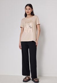 Camiseta de algodón beige con un estampado gráfico metálico en la parte frontal, combinada con pantalones negros de corte holgado y sandalias planas oscuras.
