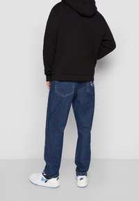 Sudadera negra de corte holgado, combinada con jeans de mezclilla azul sueltos. Zapatillas blancas con detalles en negro y azul completan el conjunto.