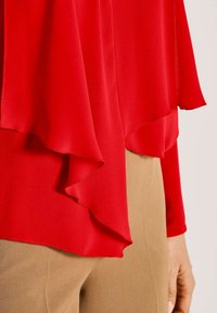 Rotes, geschichtetes Chiffon-Oberteil mit asymmetrischem Saum, getragen über beigen Hosen, mit einer Nahaufnahme der Taille und der rechten Hand.