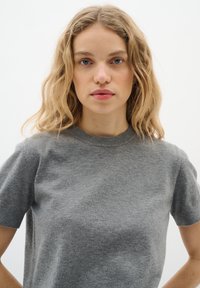 Blonde Frau mit welligem Haar, die ein kurzärmliges graues Strickoberteil trägt, blickt nach vorne vor einem schlichten weißen Hintergrund.