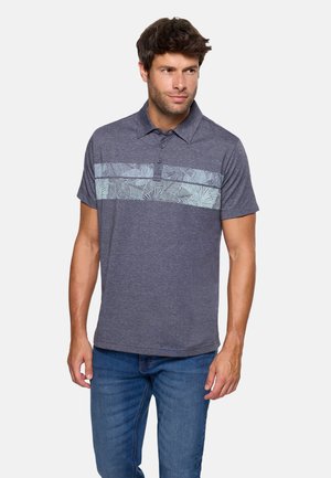 Hombre con camiseta polo de manga corta azul con patrones de hojas de palma en azul claro en el pecho y pantalones vaqueros azules, de pie contra un fondo blanco.