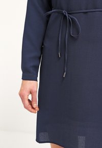 Vestido azul marino hecho de material texturizado, con mangas largas, cintura atada y detalles metálicos en el cordón.