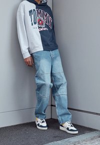 Grijze en marineblauwe hooded sweatshirt met het "TOMMY"-logo, lichtblauwe wijde spijkerbroek met naden, en witte sneakers met marineblauwe accenten en gele details.