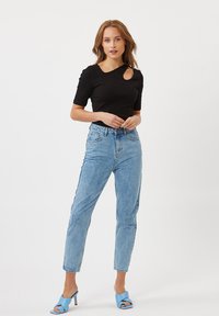 Top corto nero a maniche corte con un taglio laterale, abbinato a jeans a vita alta azzurri e sandali con tacchi blu, su uno sfondo bianco.