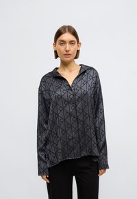 Cămașă gri cu nasturi, dintr-un material lucios, cu un model geometric de cercuri și linii. Are guler și mâneci lungi, croială lejeră.