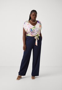 Blouse à imprimé floral avec des manches courtes et un détail de taille nouée, associée à un pantalon bleu marine à coupe fluide. Tissu texturé et sandales beiges plates.