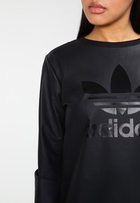 Czarna koszula z długim rękawem wykonana z gładkiego materiału, z błyszczącym czarnym logo Adidas z trzema liśćmi na przodzie.