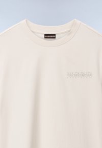 T-shirt léger beige clair à manches courtes en coton. Il présente un col rond et un logo en relief subtil sur le côté gauche de la poitrine.