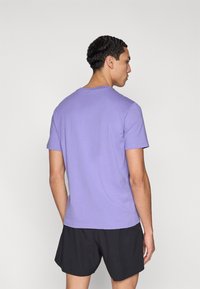 Μωβ βαμβακερό μπλουζάκι με κοντά μανίκια, απλό στρογγυλό λαιμό και απλό σχέδιο, φορεμένο με μαύρο σορτς. Πλάγια όψη από πίσω.