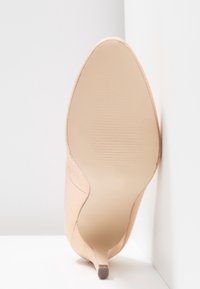 Chaussure à talon haut en daim beige à bout pointu montrée vue de la semelle, debout contre un fond blanc avec un angle.
