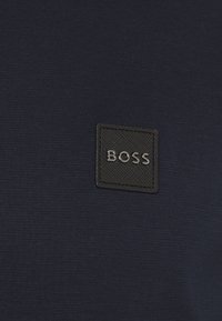 Patch carré texturé noir avec lettrage argenté "BOSS" sur fond en tissu bleu marine foncé.