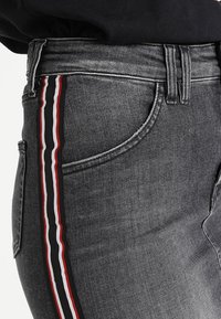 Šedé denimové džíny s úzkým černým, červeným a bílým bočním pruhom. Materiál je jemný s mírně opotřebovanou texturou.