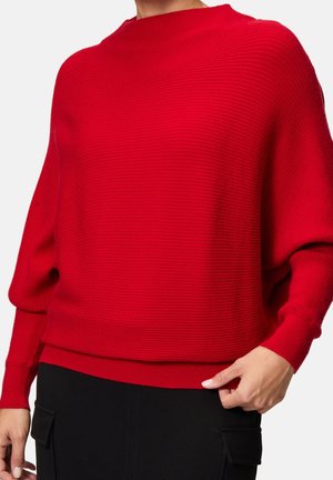 Personne portant un pull côtelé rouge vif à manches longues et un pantalon noir, main reposant près de la poche.
