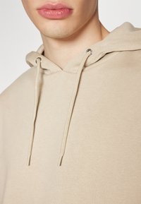 Beige Kapuzenpullover mit weicher Textur, ausgestattet mit einer Kordelzugkapuze und metallischen Ösen zur Verstärkung. Nahaufnahme zeigt den glatten Stoff und die Nähte.