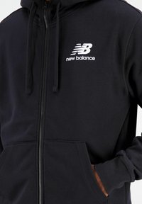 Černá mikina se zipem vyrobená z měkké látky, s klokaní kapsou, nastavitelnou kapucí se šňůrkou a bílým logem New Balance na hrudi.