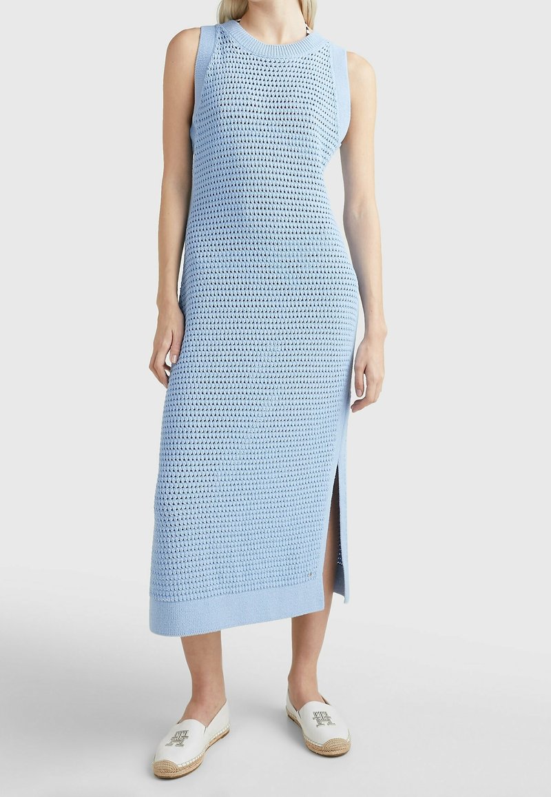 Robe midi bleu clair sans manches en maille avec fente latérale portée avec des espadrilles blanches à enfiler sur une personne debout.
