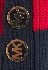 Granatowy sweter kardigan z czerwonymi akcentami, z dwoma złotymi okrągłymi guzikami z wytłoczonym logo "MK". Materiał o fakturze prążkowanej.