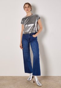 Silberner Pailletten-Crop-Top mit kurzen Ärmeln, kombiniert mit hochgeschnittenen blauen Schlagjeans und metallisch silbernen Knöchelschuhen.