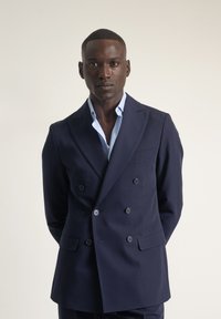 Blazer azul marino de doble botonadura con solapas con muesca, dos bolsillos frontales y botones en contraste. Lleva puesto una camisa de color azul claro.