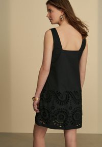 Schwarzes Kleid mit quadratischem Ausschnitt, breiten Trägern und einem detailliert bestickten Saum mit kreisförmigen Mustern. Glattes Material, knielanges Design.