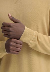 Langarmig, lichtgeel gebreide trui met een gestructureerde stof. Close-up van handen die de mouwen met geribbelde boorden vasthouden.
