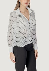Blusa bianca trasparente con pois neri, chiusura a bottoni, design plissettato, collo a punta e ampi polsini, abbinata a pantaloni neri.