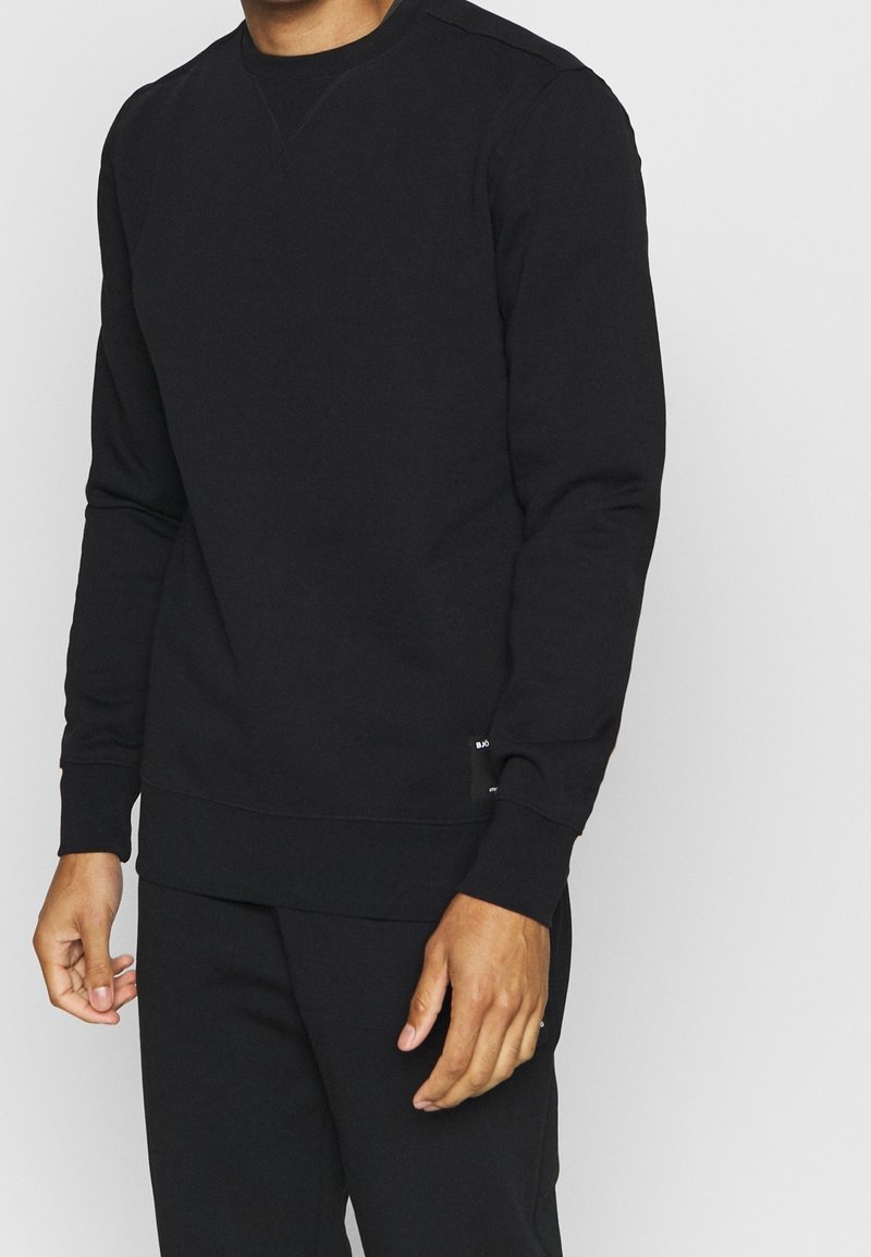 Homme portant un sweat-shirt noir uni à manches longues et un pantalon noir assorti sur un fond gris clair.