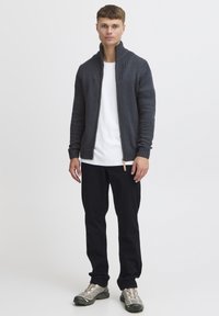 Kolettad, stickad cardigan i kolgrått med hög ribbad krage, framdragkedja och en kontrasterande vit t-shirt under. Matchad med svarta byxor och sneakers.