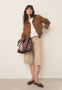 Jachetă maro din piele întoarsă, top crem, pantaloni scurți bej până la genunchi și balerini maro închis. Ținând o geantă cu imprimeu leopard și mânere.